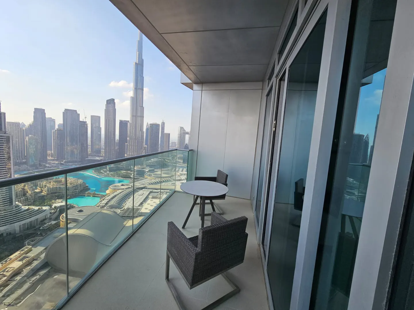 Balcony view of Dubai skyline with Burj Khalifa. Table and chairs on balcony with glass railing. Sliding glass doors to the right.