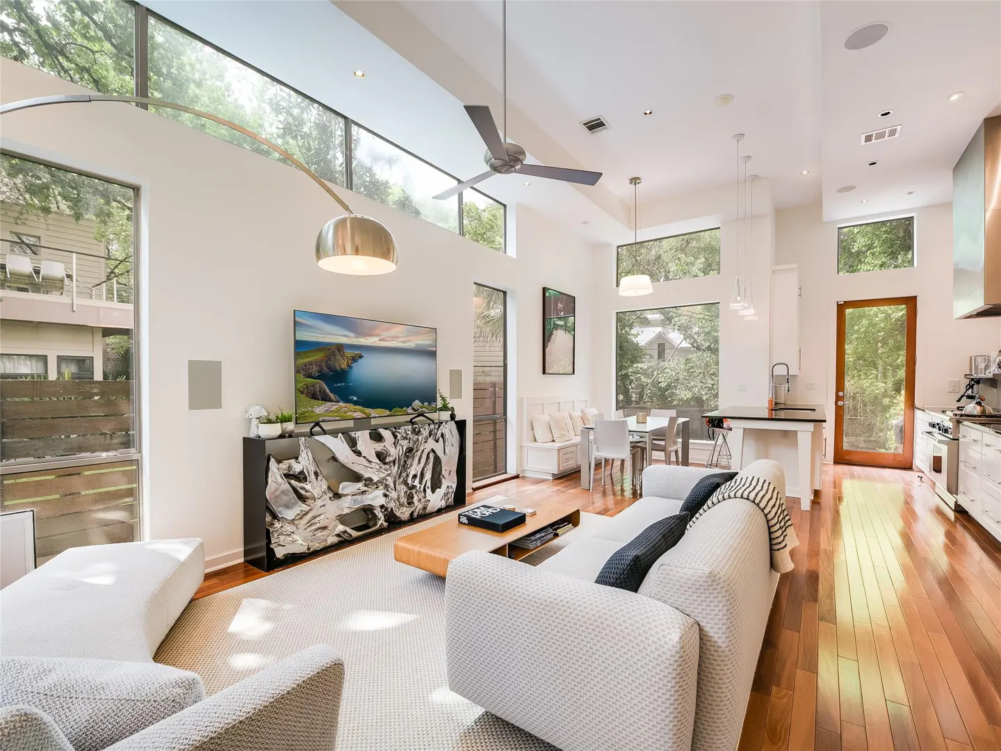 Bright, open-concept living room with white walls, wood floors, and large windows. A white sofa and chairs sit on a light rug. A TV displays a coastal scene.