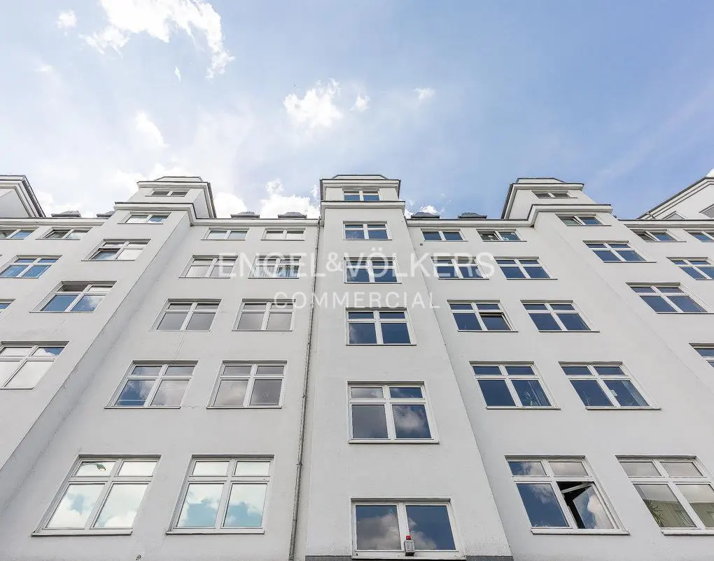 Low angle view of a white commercial building under a blue sky with clouds. Windows reflect the sky. "Engel & Völkers Commercial" logo is superimposed.