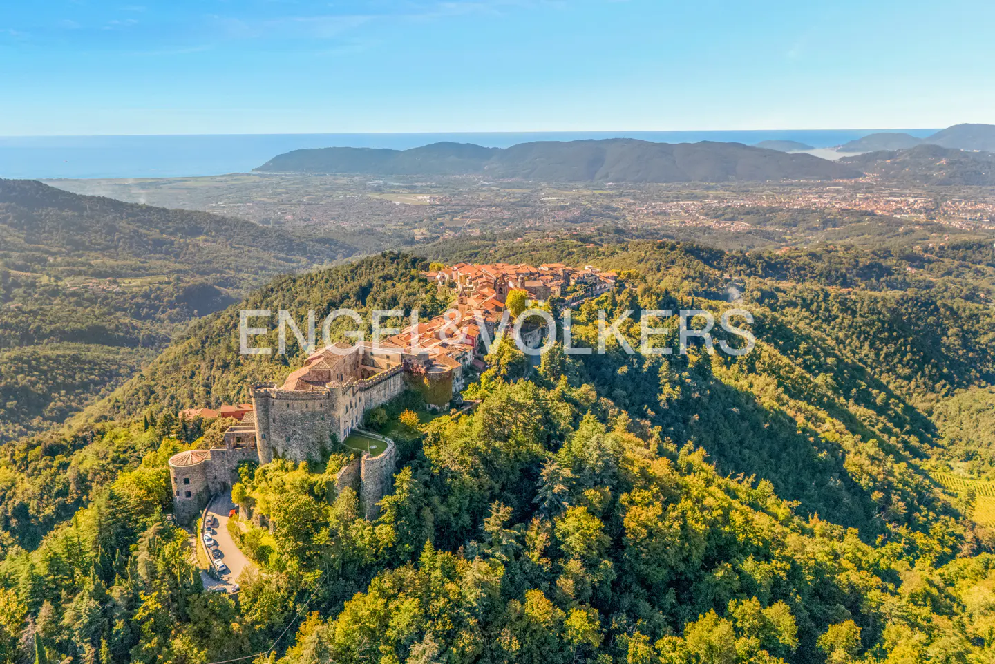 Aerial view of a stone castle and village atop a green, tree-covered hill, with a city and ocean in the background under a blue sky.
