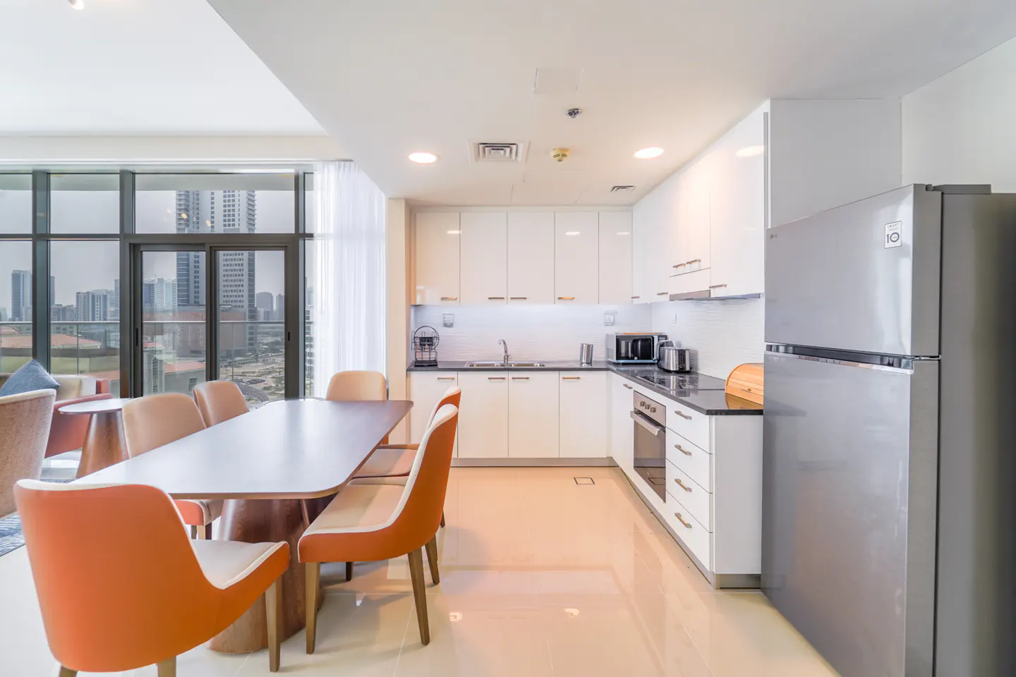 Bright, modern kitchen with white cabinets, stainless steel appliances, and orange chairs around a dining table. City view through large windows.
