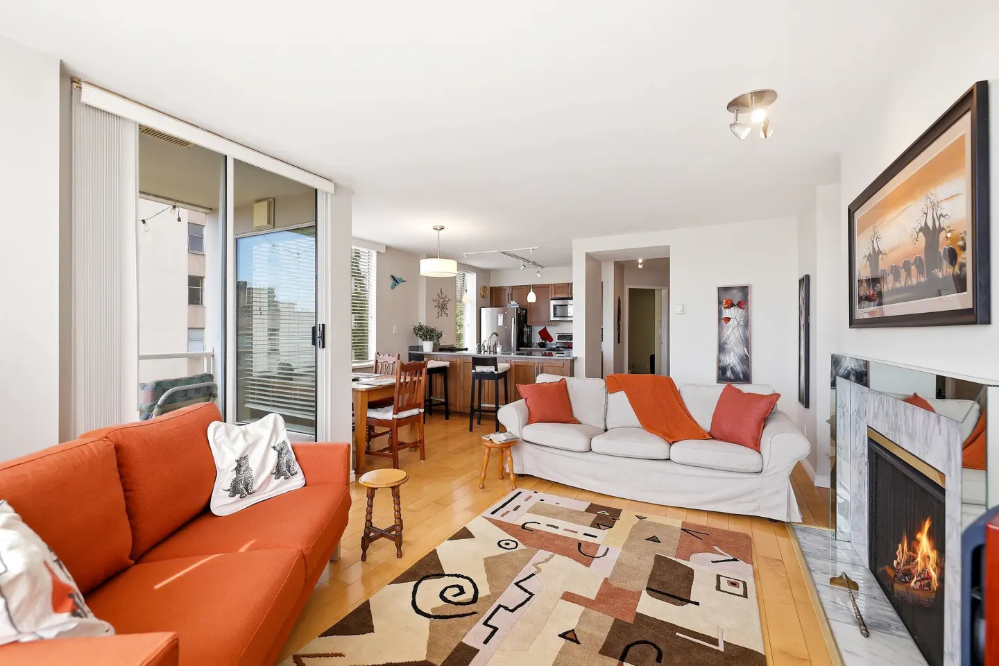 Bright living room with orange sofa, white sofa, patterned rug, and fireplace. Kitchen and dining area in background.
