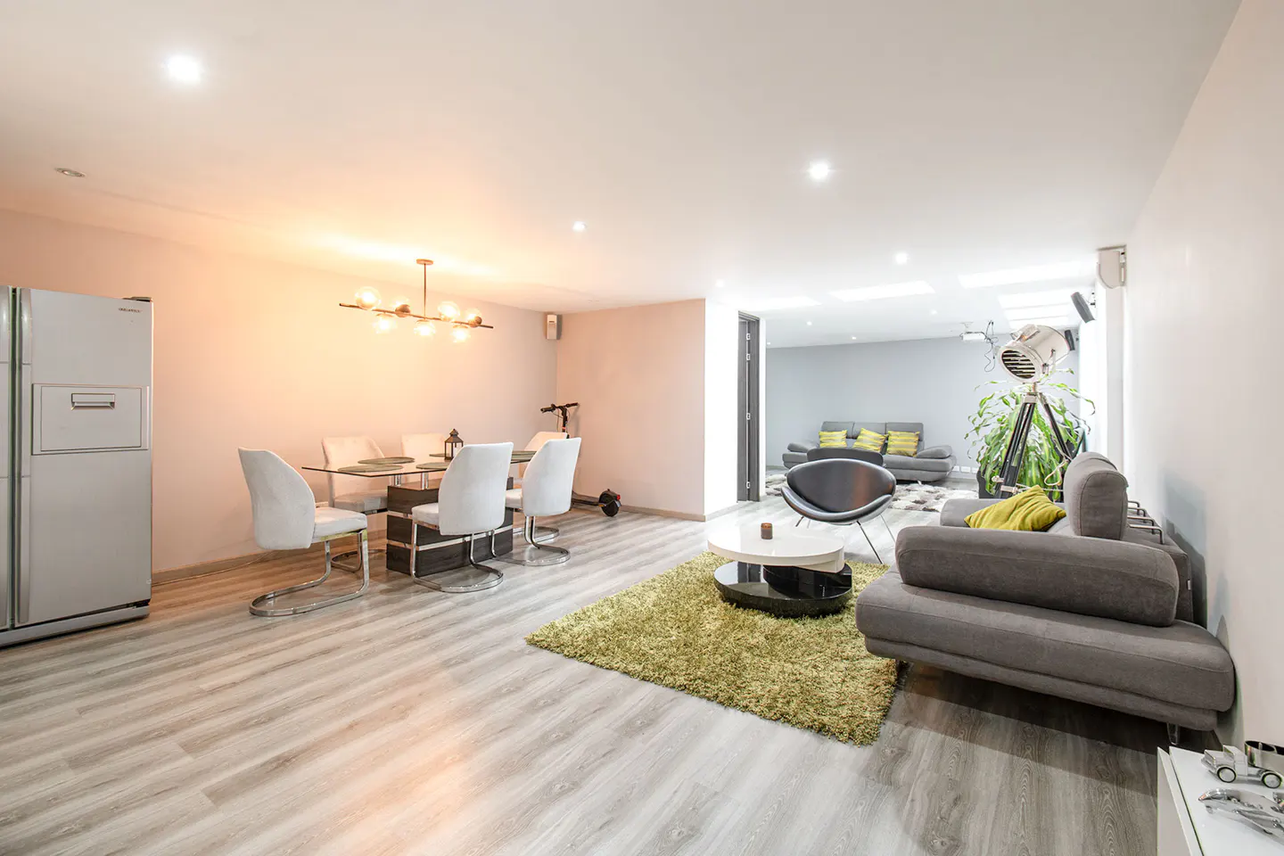 Bright, open-concept living and dining area with gray wood floors, gray sofa, and glass dining table with white chairs. A large silver refrigerator is on the left.