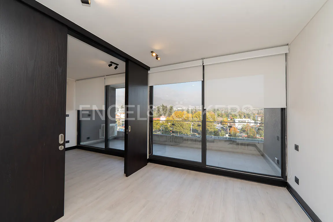 A bright, empty room with light wood floors and a large window with white blinds overlooking a city. A dark wood sliding door is partially open.