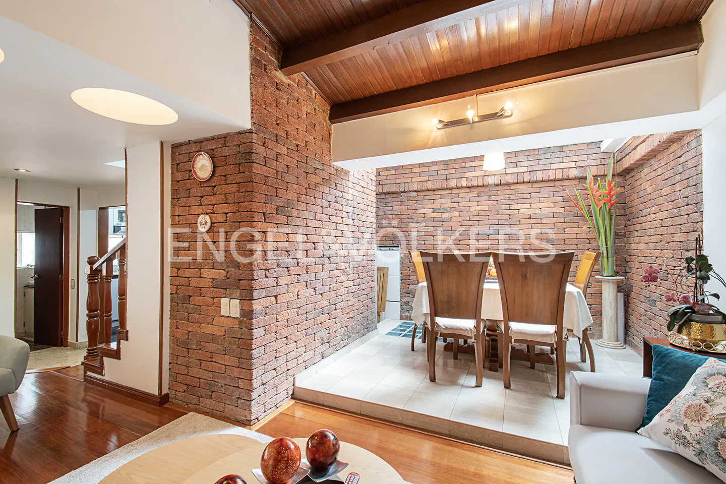 A dining area with brick walls and wood ceiling. Table with chairs, flowers, and a sofa with pillows are visible.
