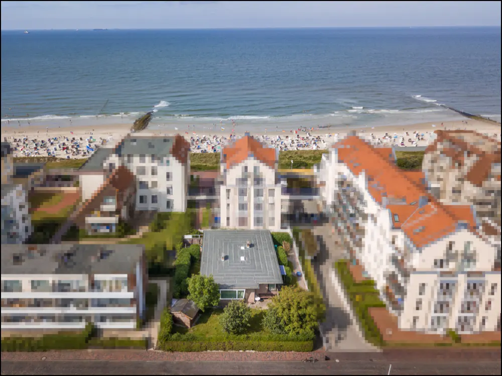 Aerial view of beachfront property with white buildings, orange roofs, and a sandy beach with people and blue ocean.