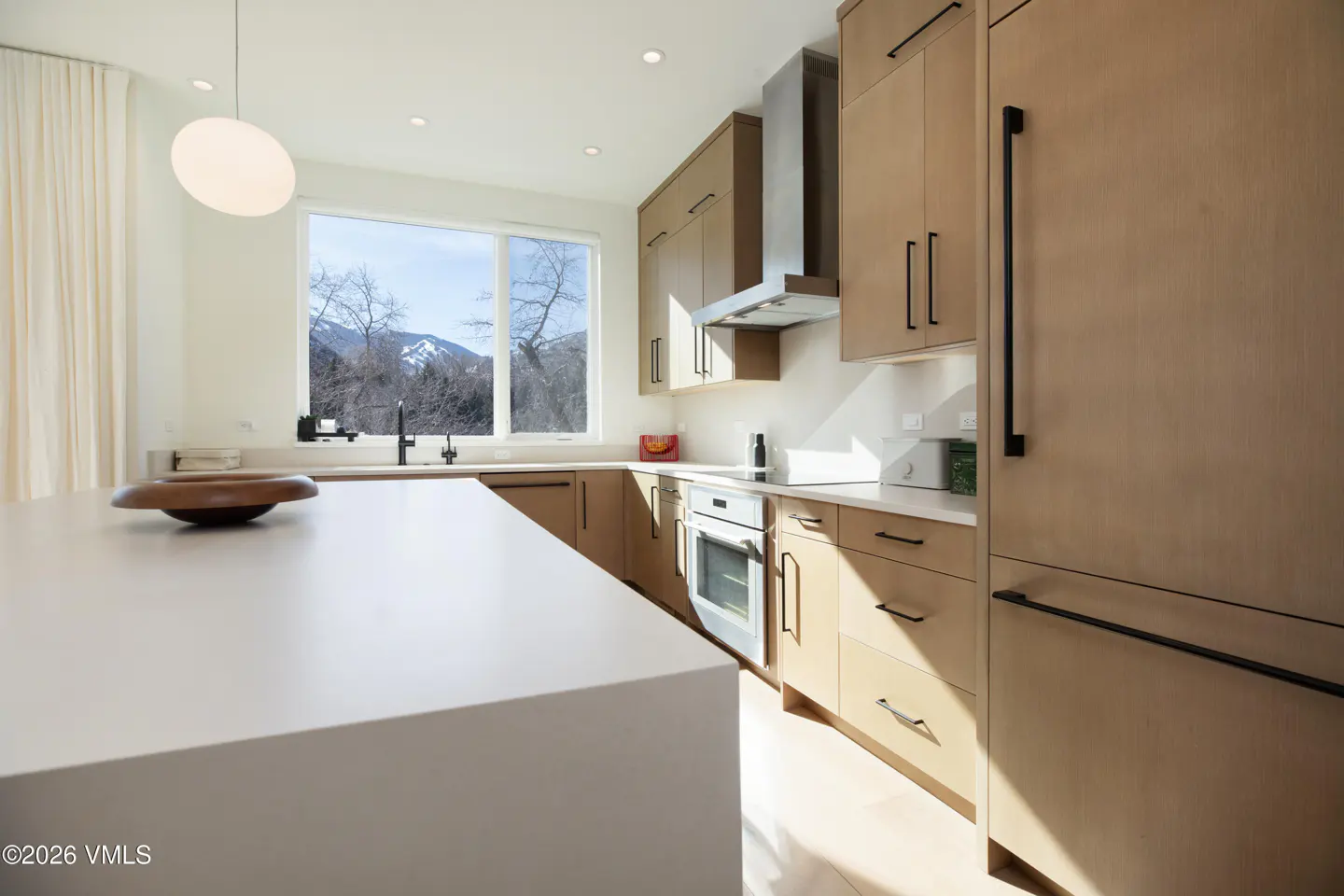 Bright kitchen with light wood cabinets, white countertops, and a large window with mountain views. A white pendant light hangs above the island.