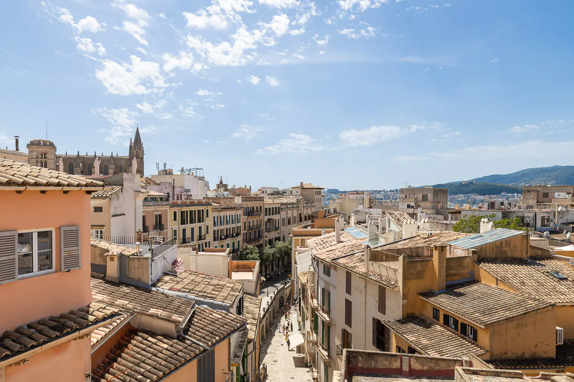 Aerial view of Palma de Mallorca, Spain, featuring terracotta rooftops, narrow streets, and a distant cathedral under a blue sky.