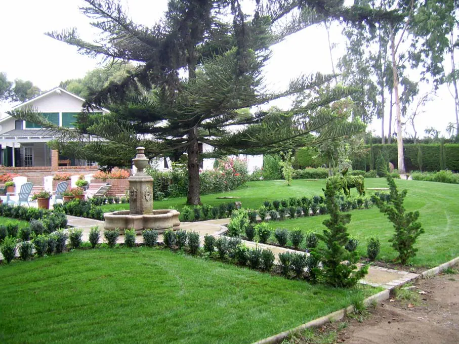 A lush green lawn with a stone fountain in the foreground and a white house in the background.