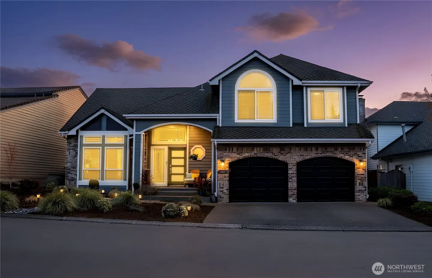Two-story home with blue siding, brick accents, and two black garage doors at dusk with warm interior lighting.