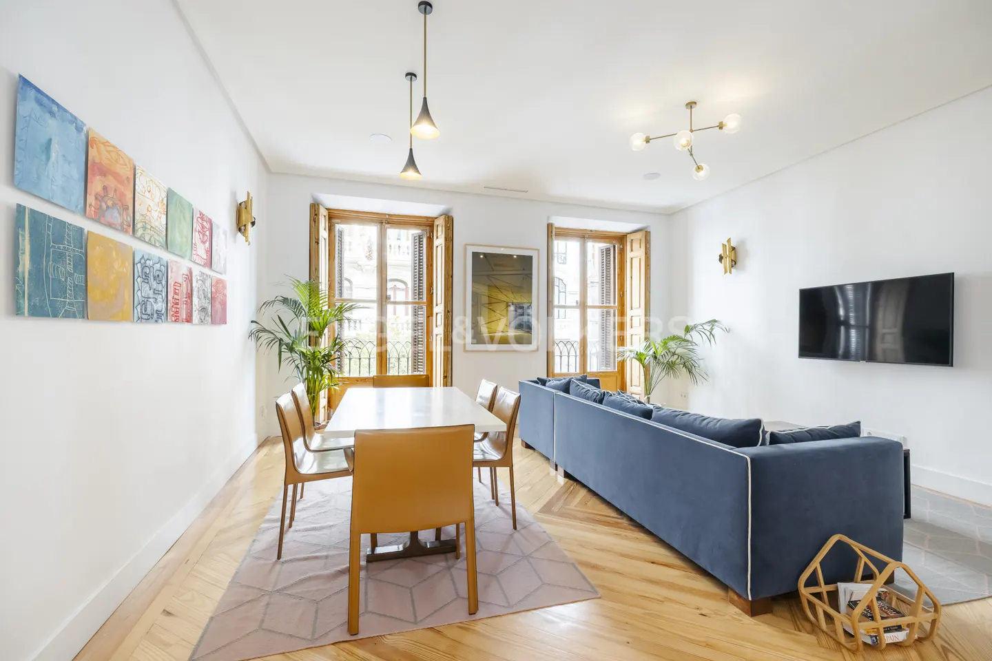 Bright living room with wood floors, white walls, and large windows. A blue sofa faces a TV, and a dining table sits under pendant lights. Artwork adorns the walls.