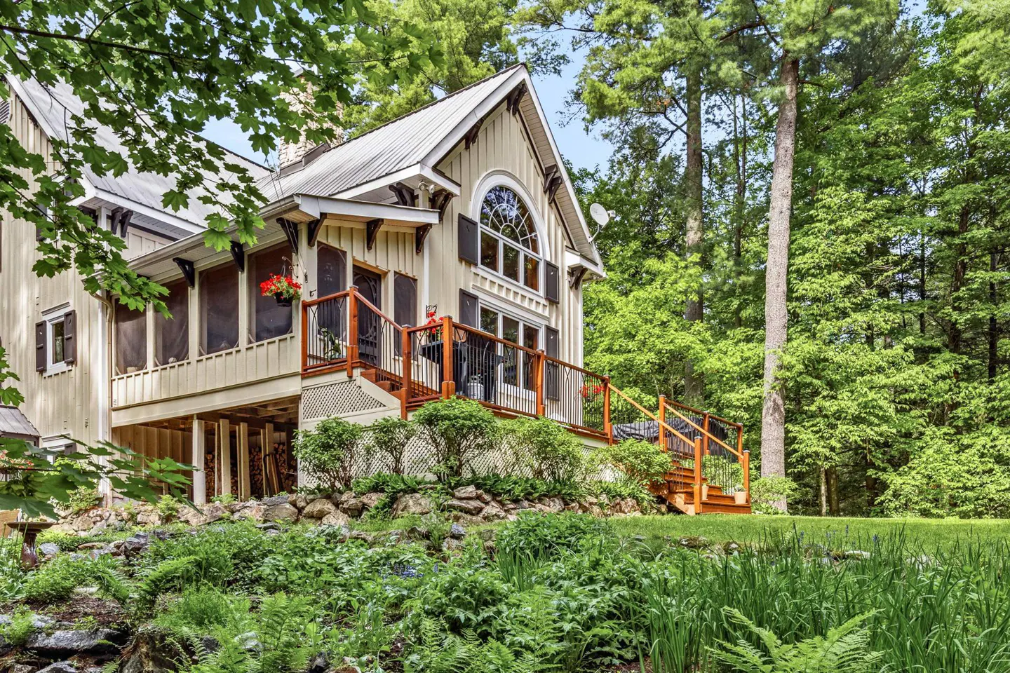 Cream-colored cottage with a metal roof, wooden deck, and arched window, nestled in a lush, green forest setting.