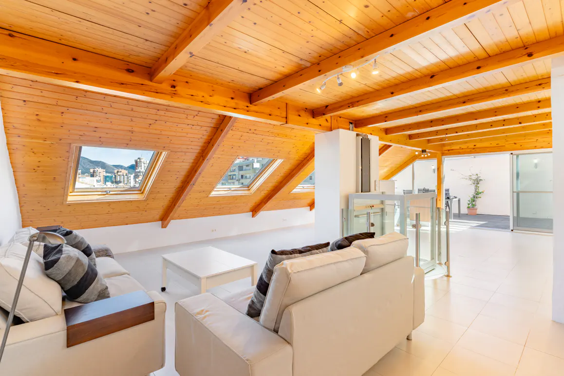 Attic living room with wood-paneled vaulted ceiling, white sofas, skylights, and a glass elevator leading to a terrace.