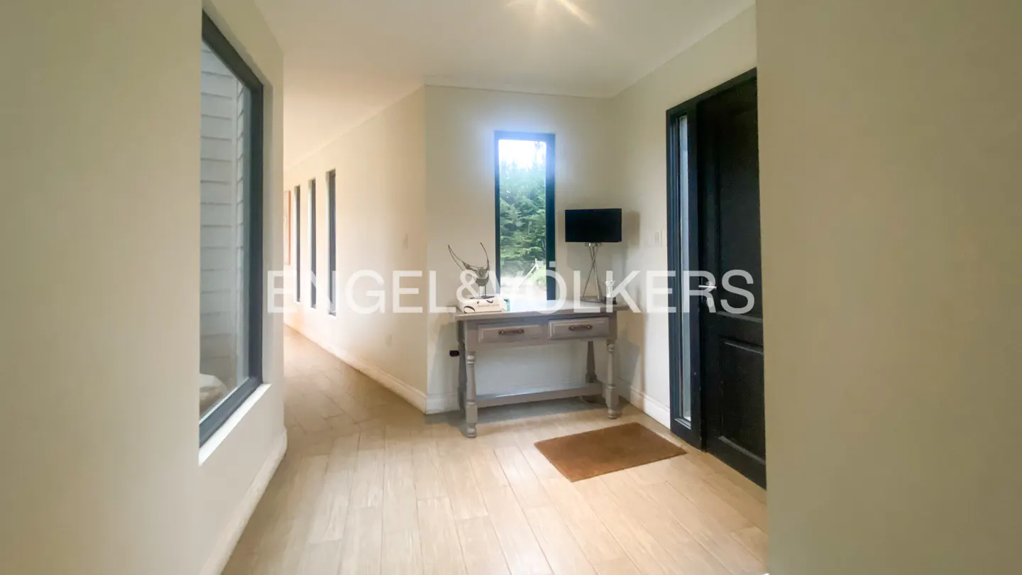Hallway with light wood floors, beige walls, and a dark door. A console table sits under a tall window with a lamp and decor.