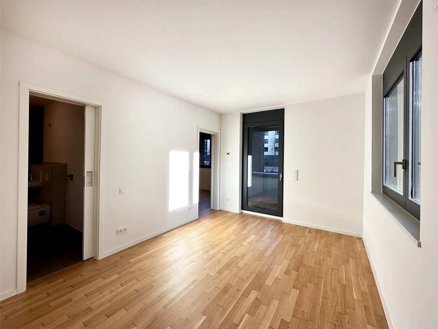 Bright, empty room with light wood floors, white walls, a dark-framed balcony door, and a doorway leading to a bathroom.