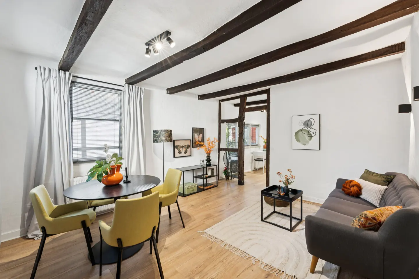 Living room with exposed beams, a gray sofa, yellow chairs, and a black table. A doorway leads to another room.