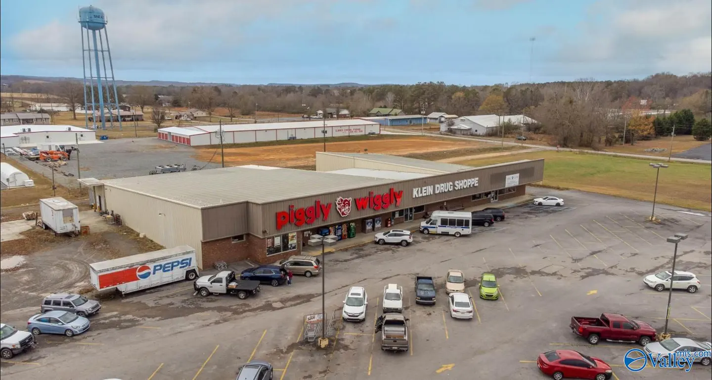 Aerial view of a Piggly Wiggly grocery store and Klein Drug Shoppe with a full parking lot and a water tower in the background.