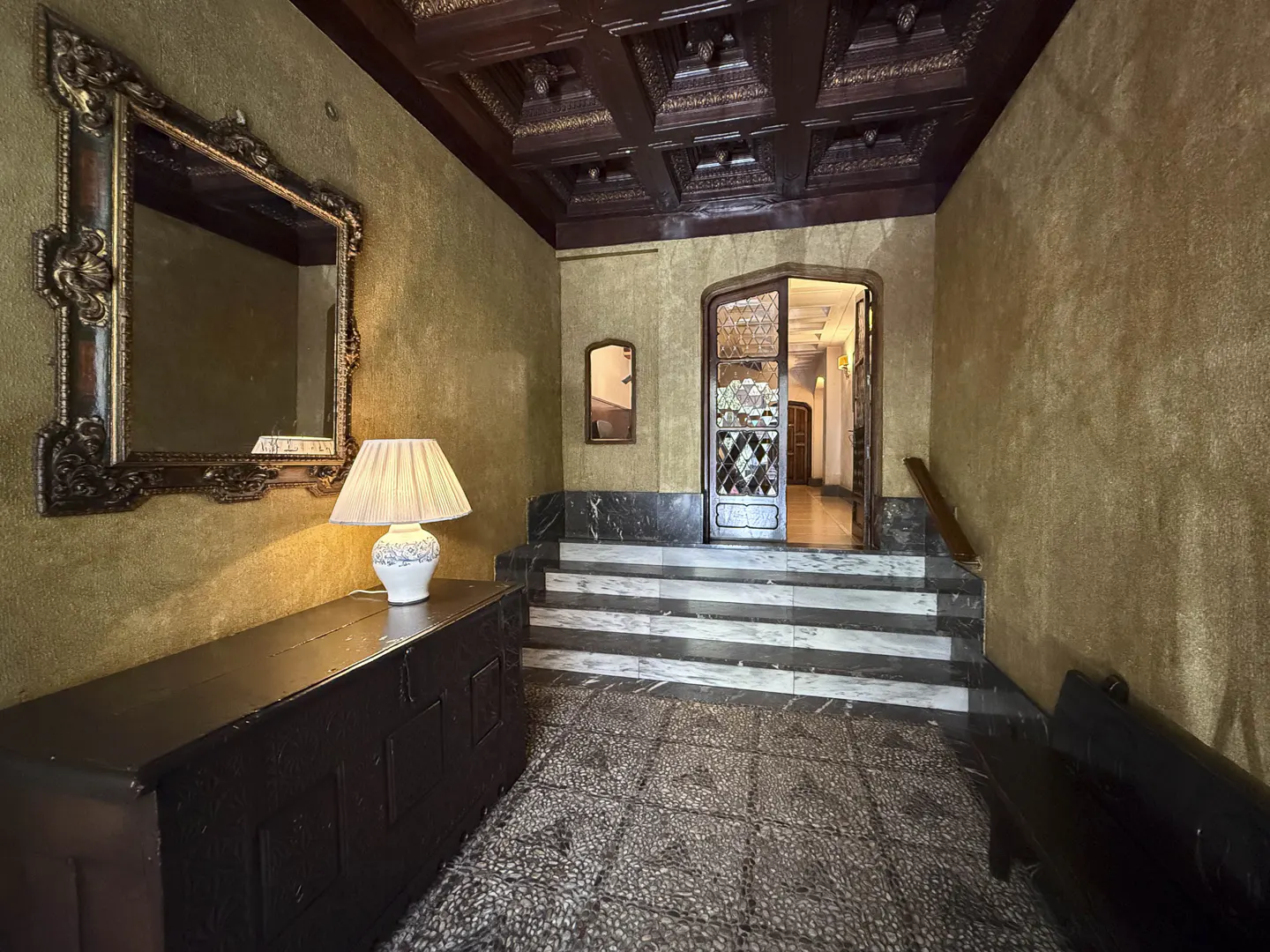 Elegant foyer with gold walls, dark wood ceiling, and stone floor. A large mirror and lamp sit atop a dark wood chest. Stairs lead to a stained glass door.