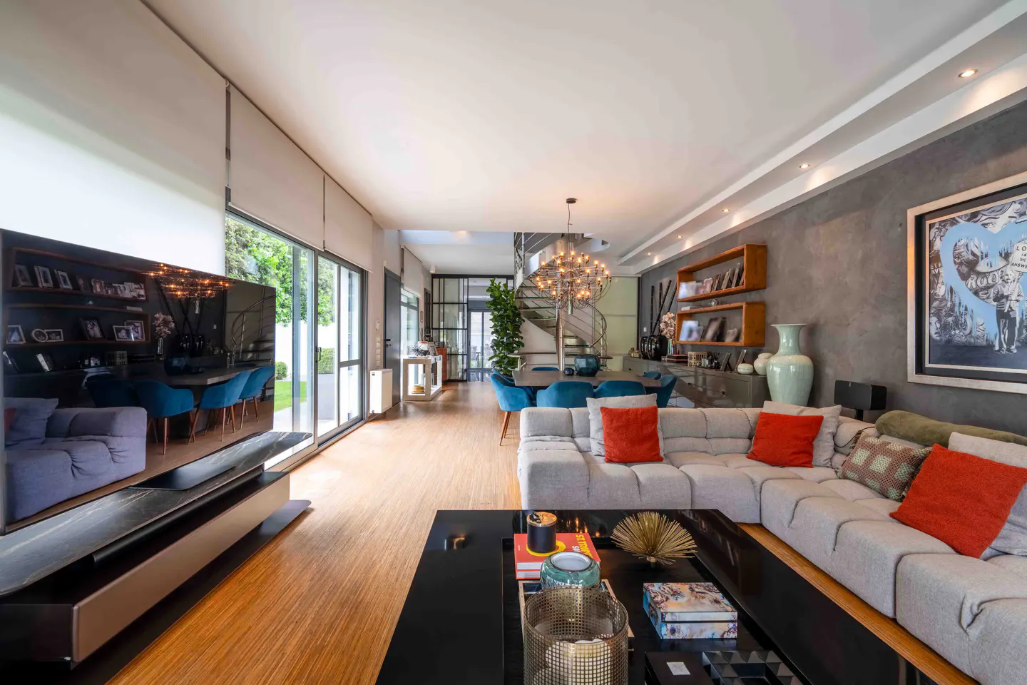 Open-concept living room with gray sectional, orange pillows, black coffee table, and wood floors. Dining area with blue chairs and spiral staircase.
