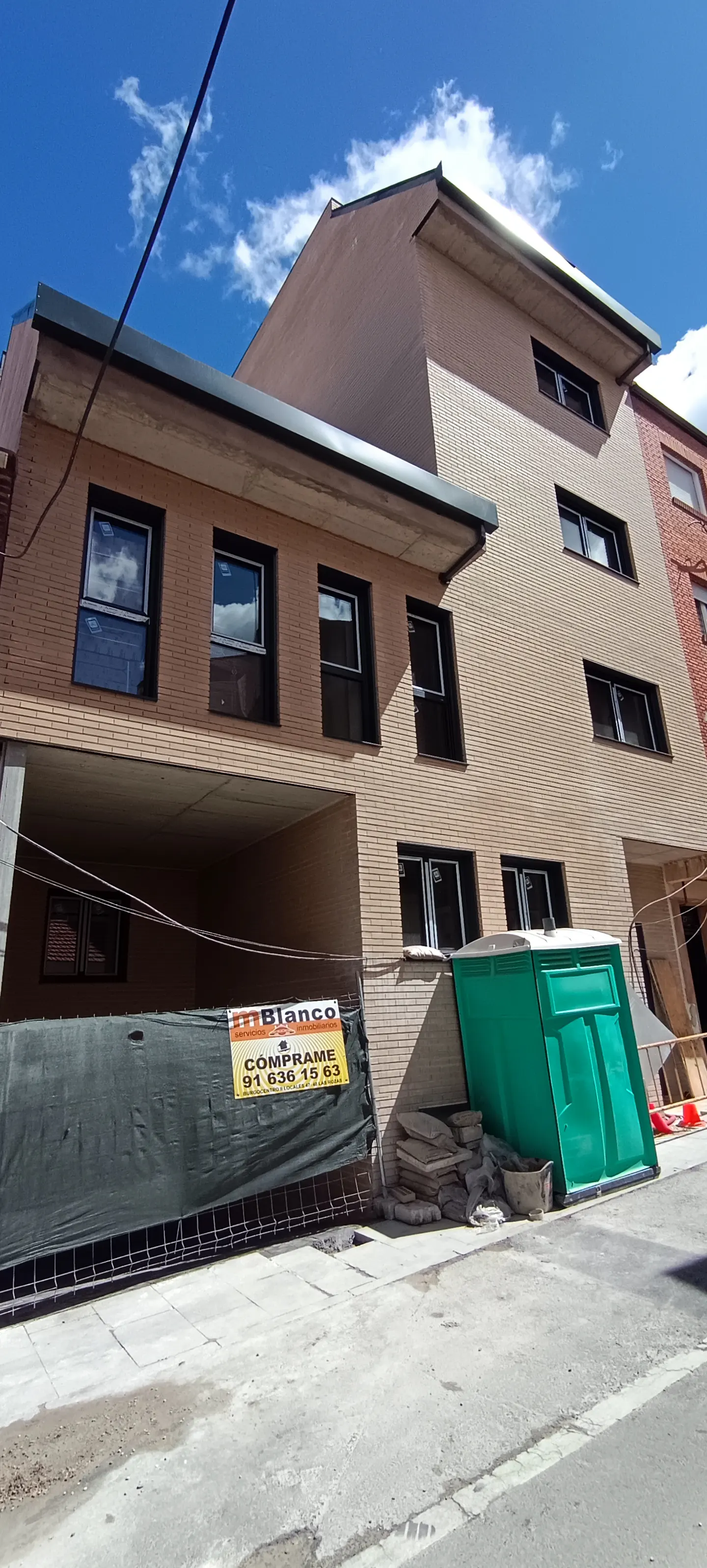 A tan brick building with black framed windows under a blue sky. A green portable toilet sits on the sidewalk in front of the building.