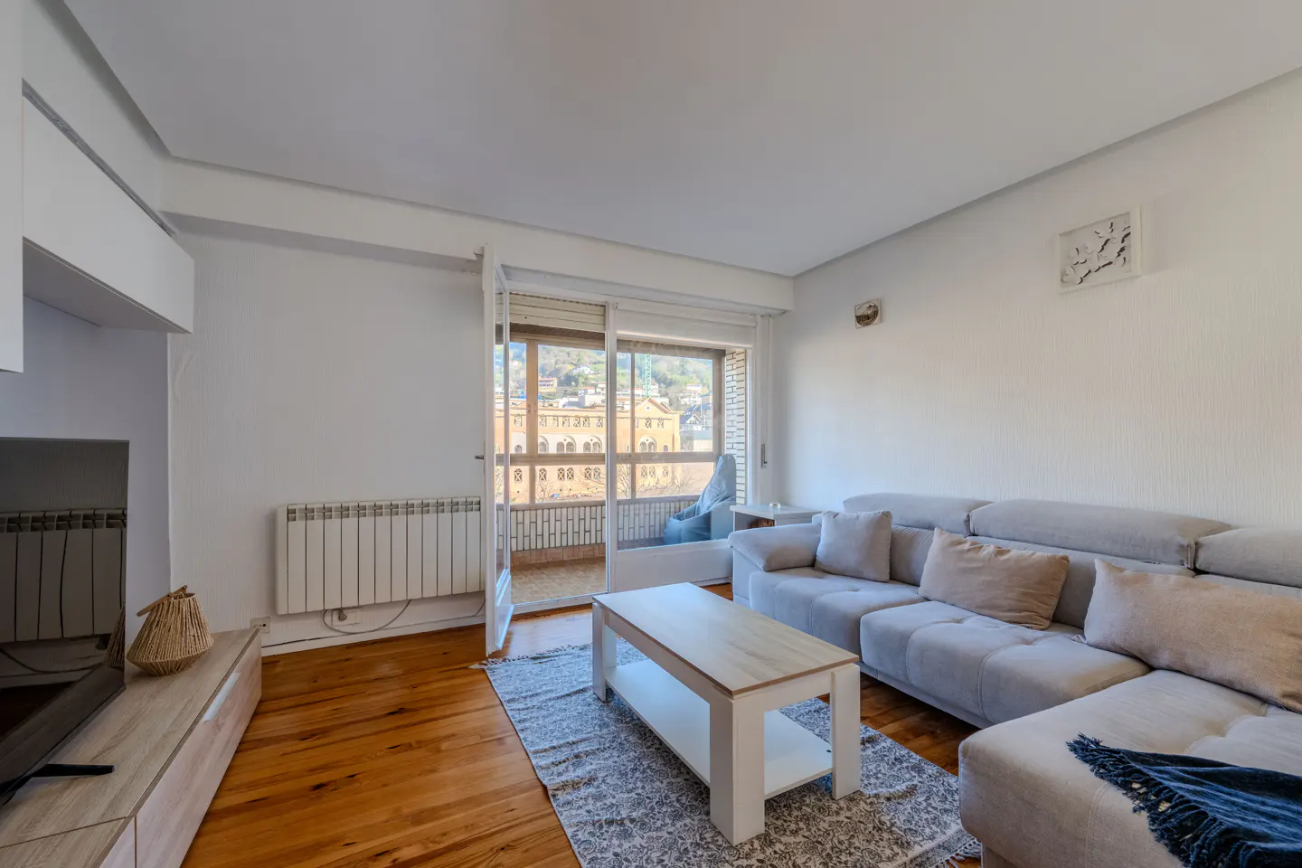 Bright living room with hardwood floors, a gray sectional sofa, and a white coffee table on a patterned rug. Balcony doors open to a city view.