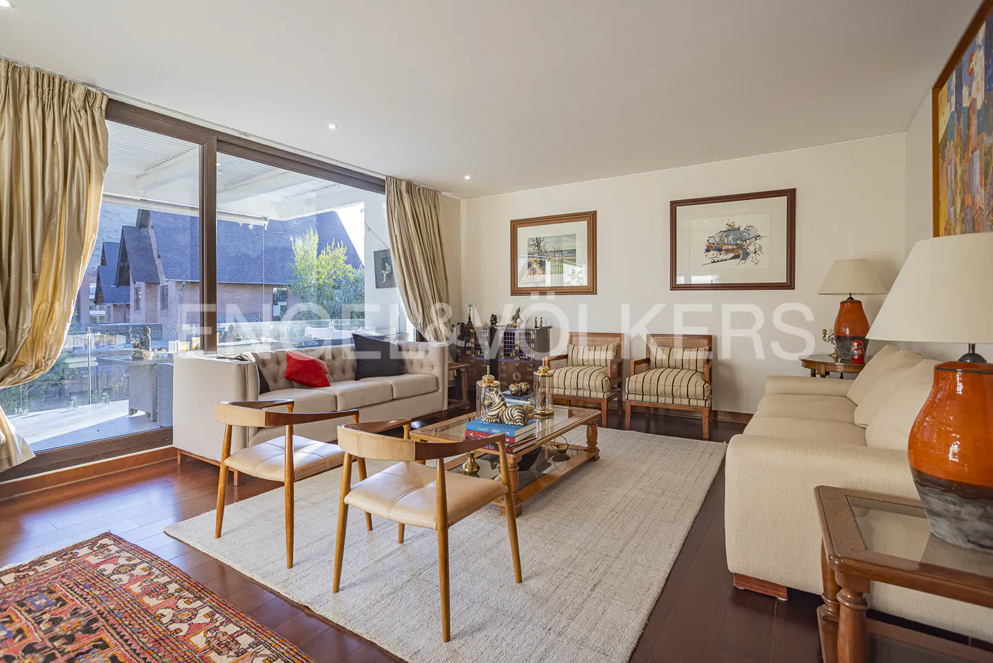 Living room with beige sofas, wooden chairs, and a glass coffee table on a white rug. Large windows with curtains offer a view of houses.