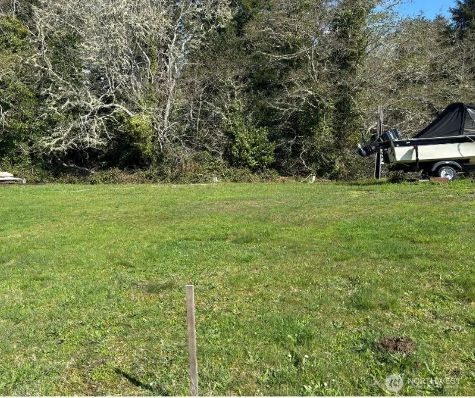A grassy lot with trees in the background and a boat on a trailer to the right.
