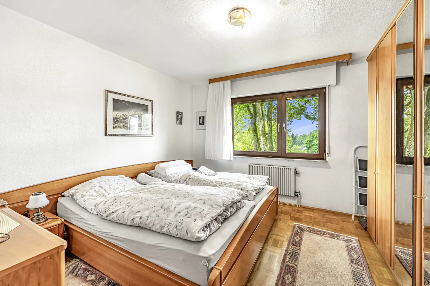 Bedroom with two twin beds, wooden frames, patterned bedding, parquet floors, and a large window overlooking green trees.