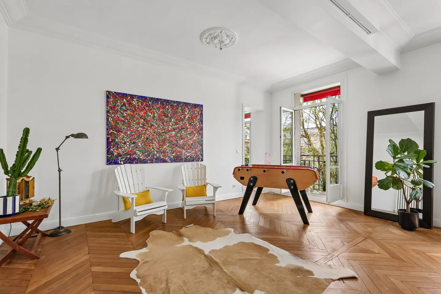 Bright living room with white walls, herringbone wood floors, and a foosball table. Two white chairs face a colorful abstract painting. A cowhide rug sits on the floor.