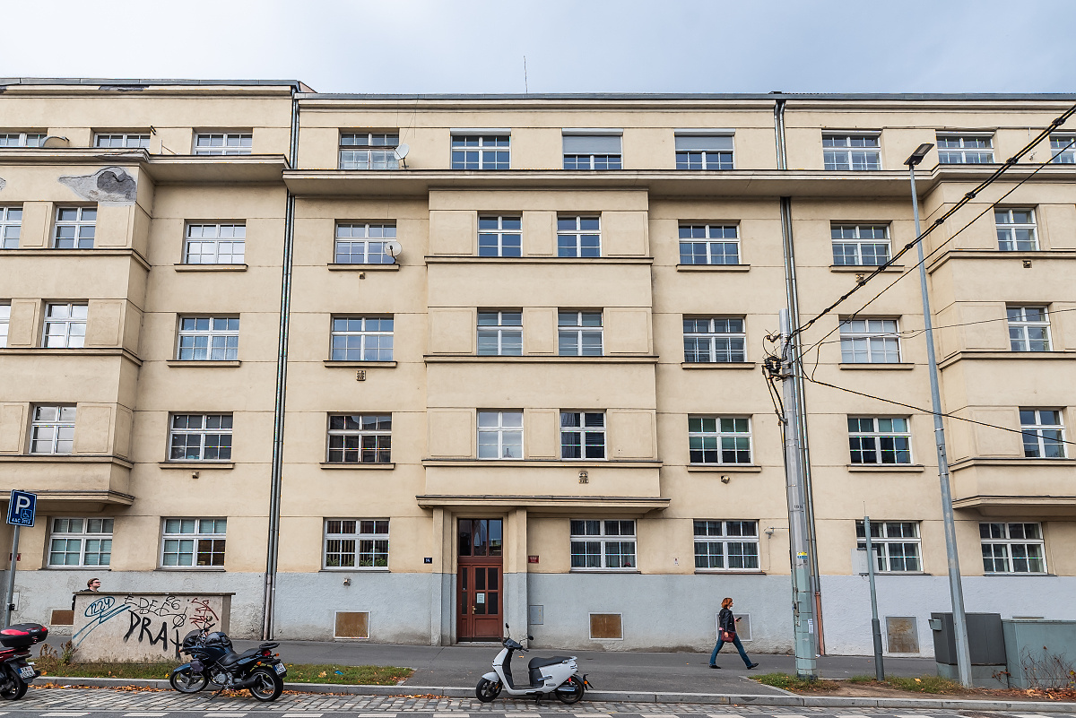 A multi-story beige apartment building with a gray base, featuring many windows, a central entrance, and parked scooters. [AI-generated text]