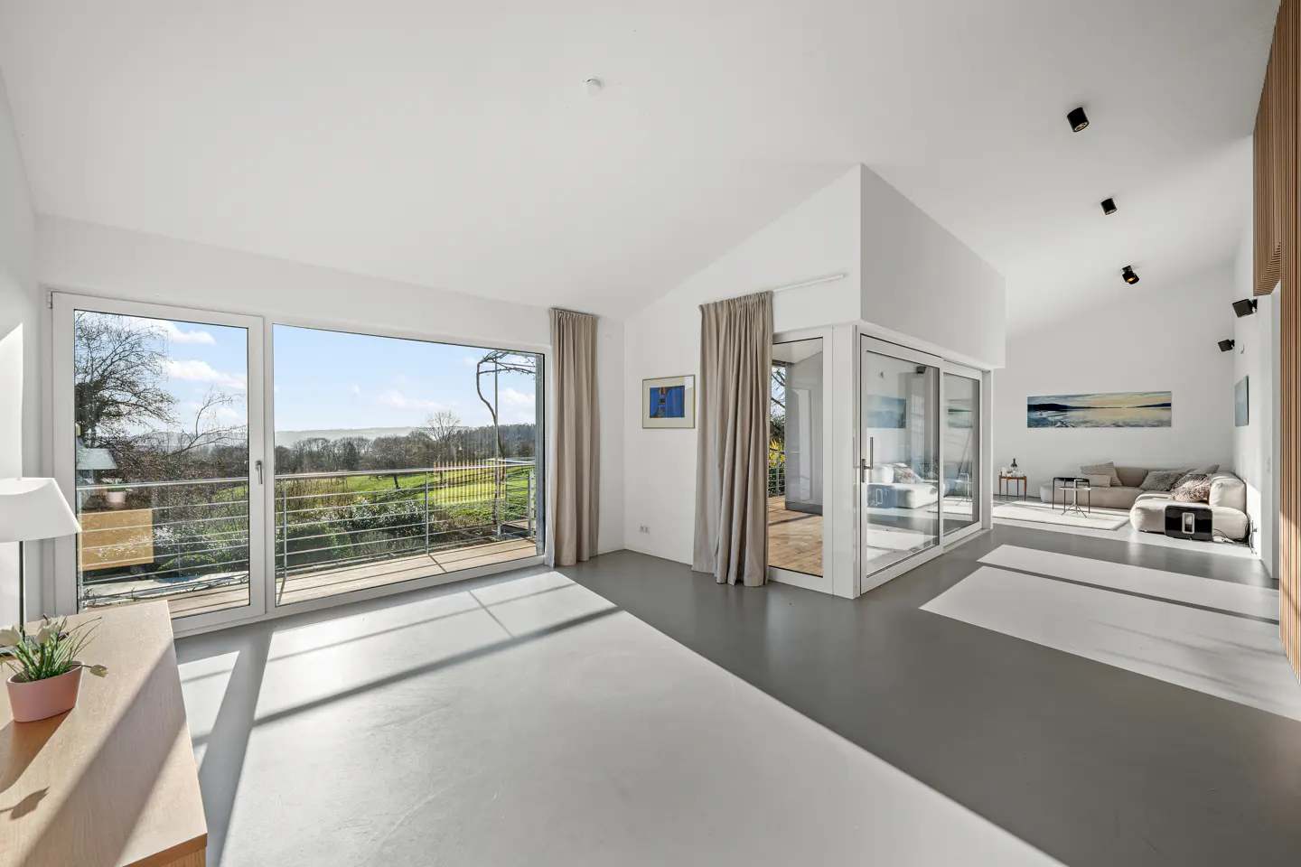 Bright, modern living room with gray floors, white walls, and large windows overlooking a green landscape. A beige sofa is visible in the background.