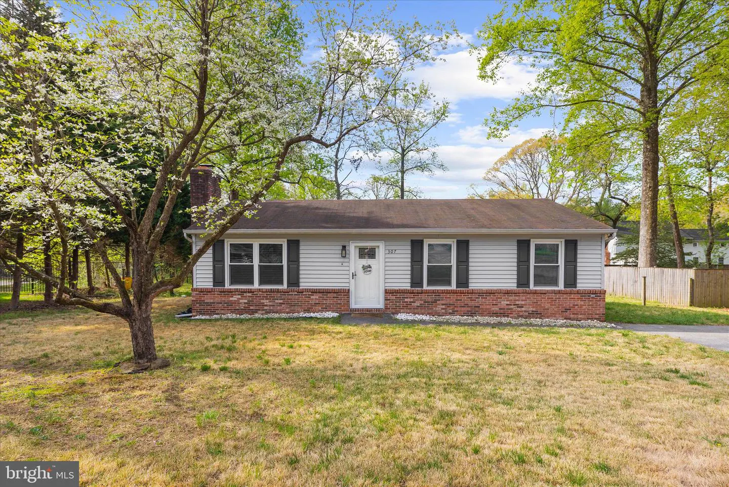 A single-story house with white siding, a brick foundation, and black shutters. A tree with white blossoms is on the left.
