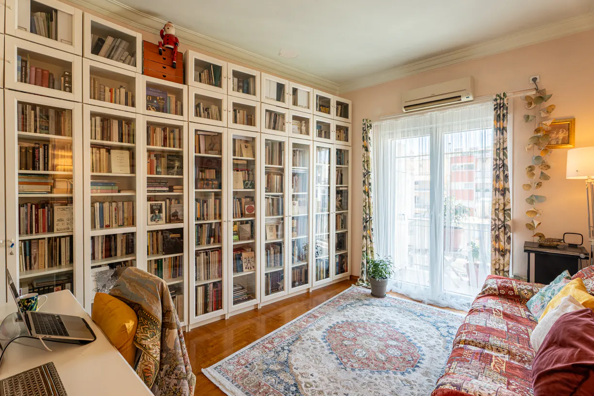 A cozy room with a large white bookcase filled with books, a desk with a laptop, and a colorful rug and sofa.