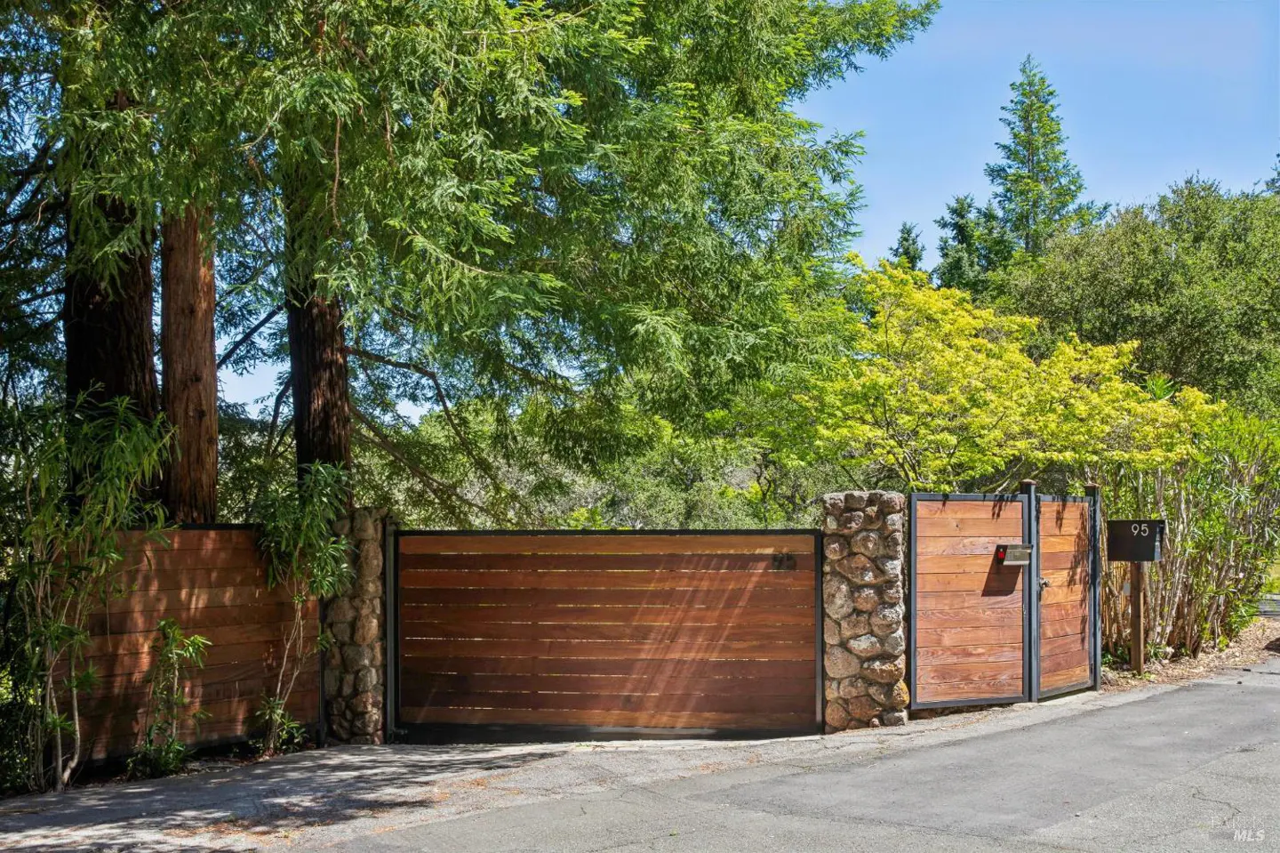 Gated entrance to a property with a wood gate, stone pillars, and lush green trees.