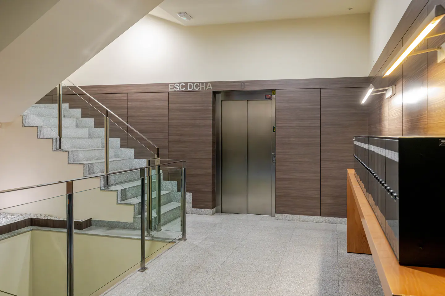 Interior view of a building lobby with stairs, elevator, and mailboxes. The stairs have glass railings and the floor is tiled.