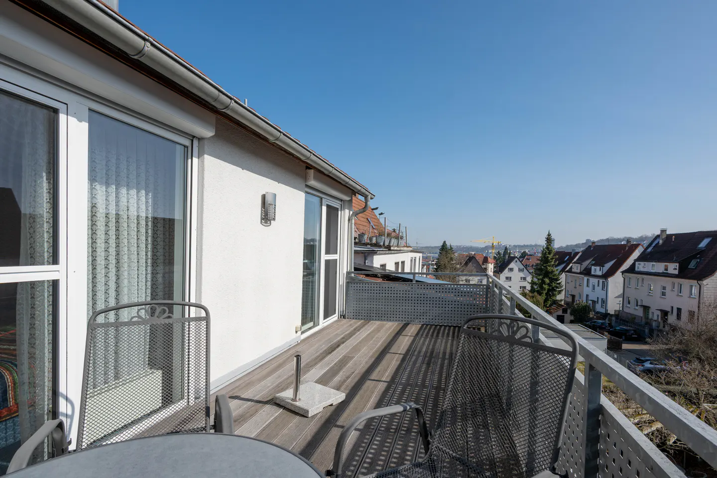 Balcony with metal chairs and table, wood-look flooring, and metal railing overlooking a neighborhood under a clear blue sky.
