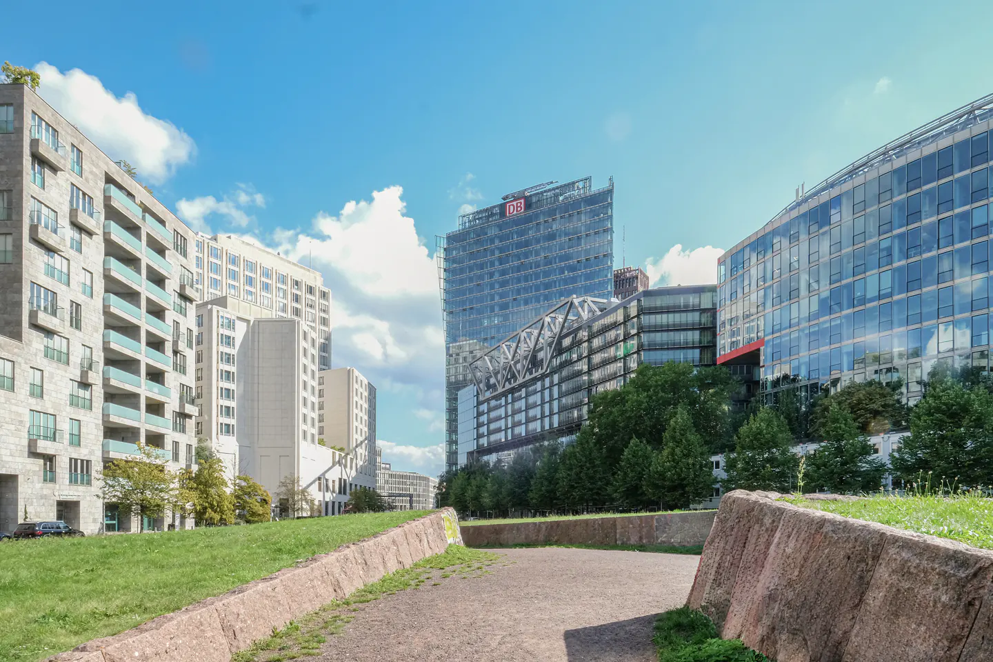 Cityscape view of modern buildings under a blue sky with white clouds. A grassy area and stone barriers are in the foreground.