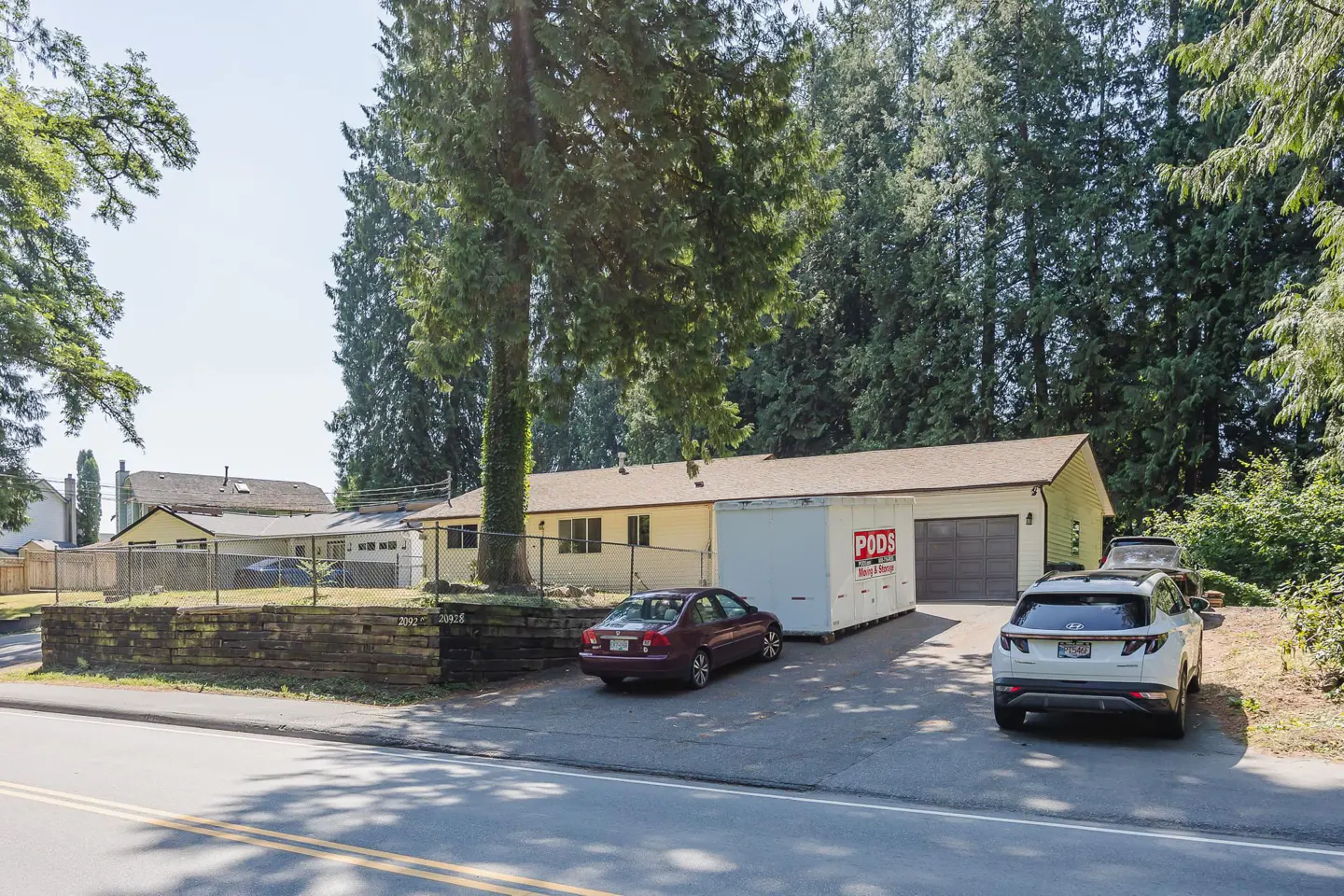 A yellow single-story house with a PODS storage container in the driveway, parked cars, and tall trees in the background.