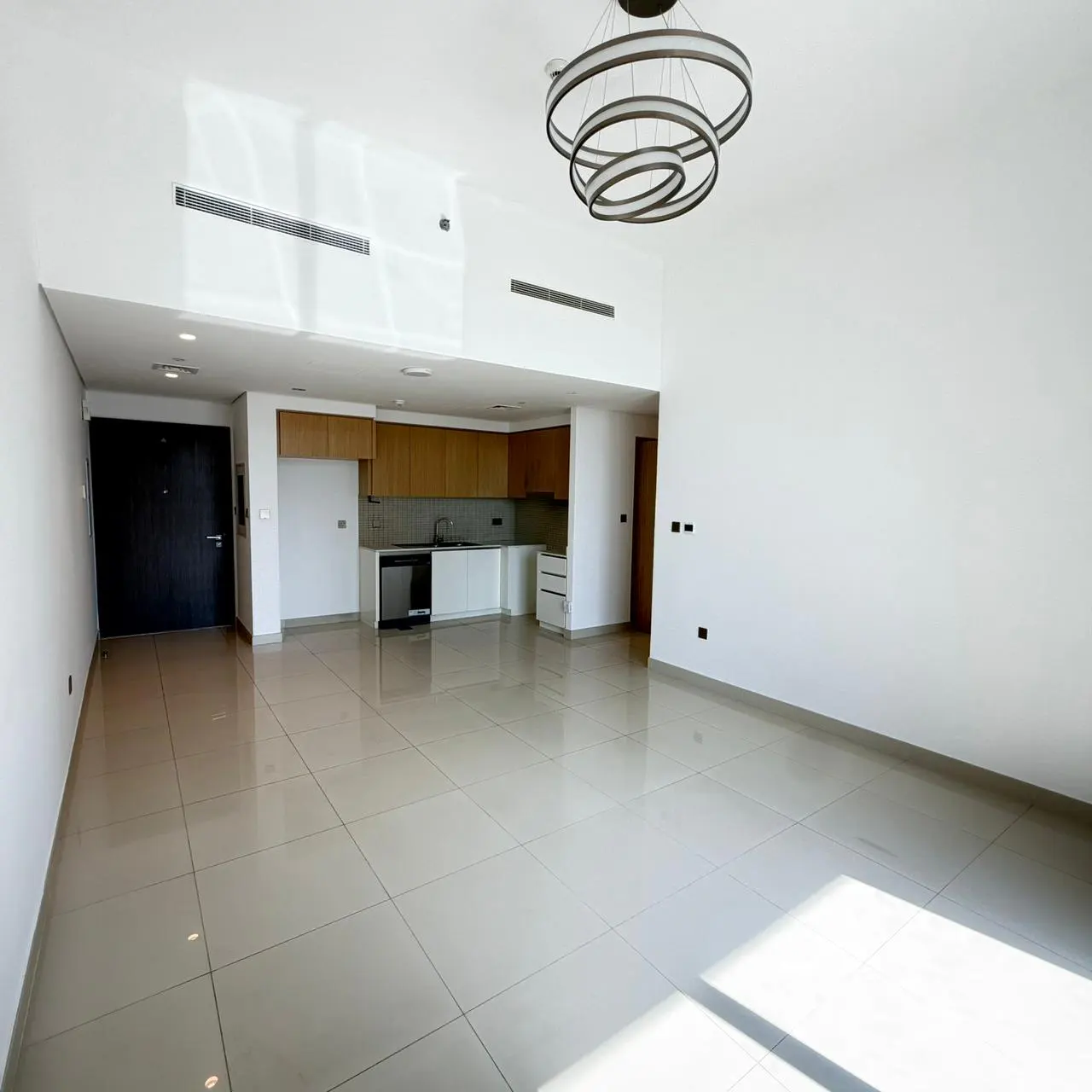 A bright, empty apartment with white walls, beige tile floors, and a modern ring chandelier. A kitchen with wood cabinets is visible in the background.