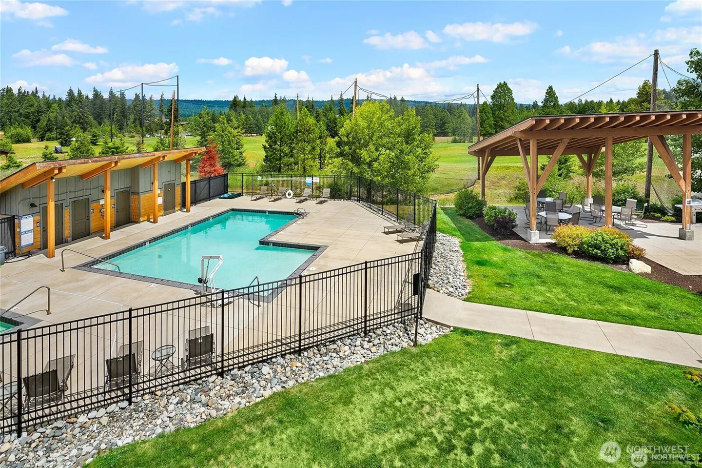 Outdoor swimming pool with a fenced deck, adjacent covered patio with tables, and a scenic view of trees and green hills.