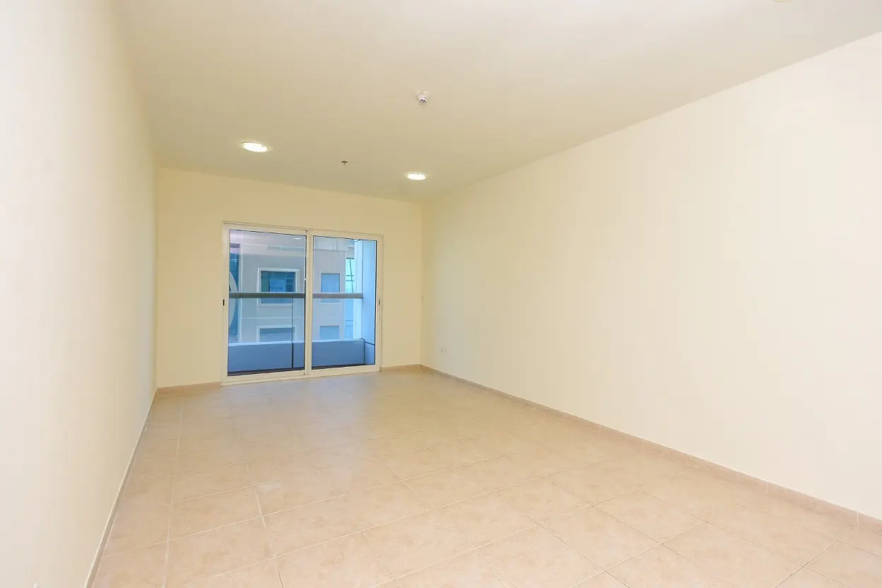 Empty room with beige walls and tiled floor. Sliding glass doors lead to a balcony with a view of a building.