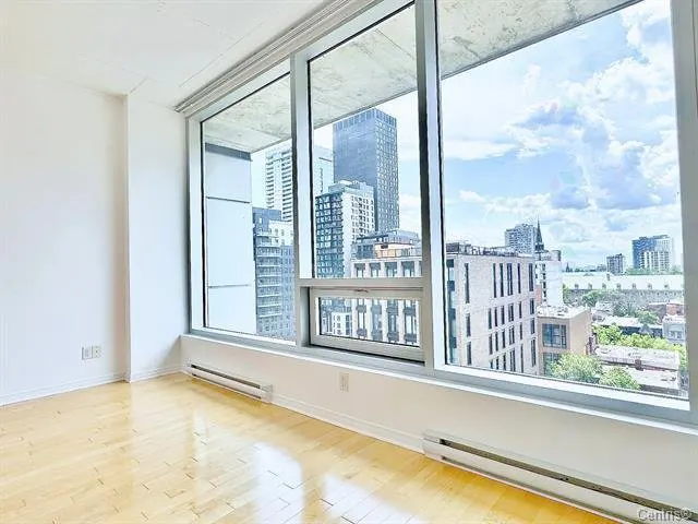 Empty room with light wood floors and a large window showing a city skyline with tall buildings.