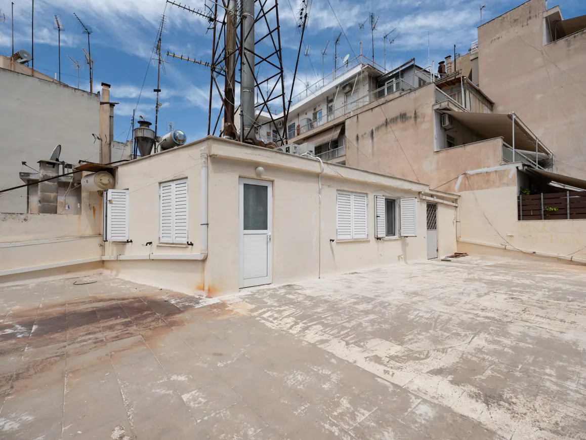 A spacious, weathered rooftop terrace with a cream-colored building featuring white shutters and a metal antenna tower.