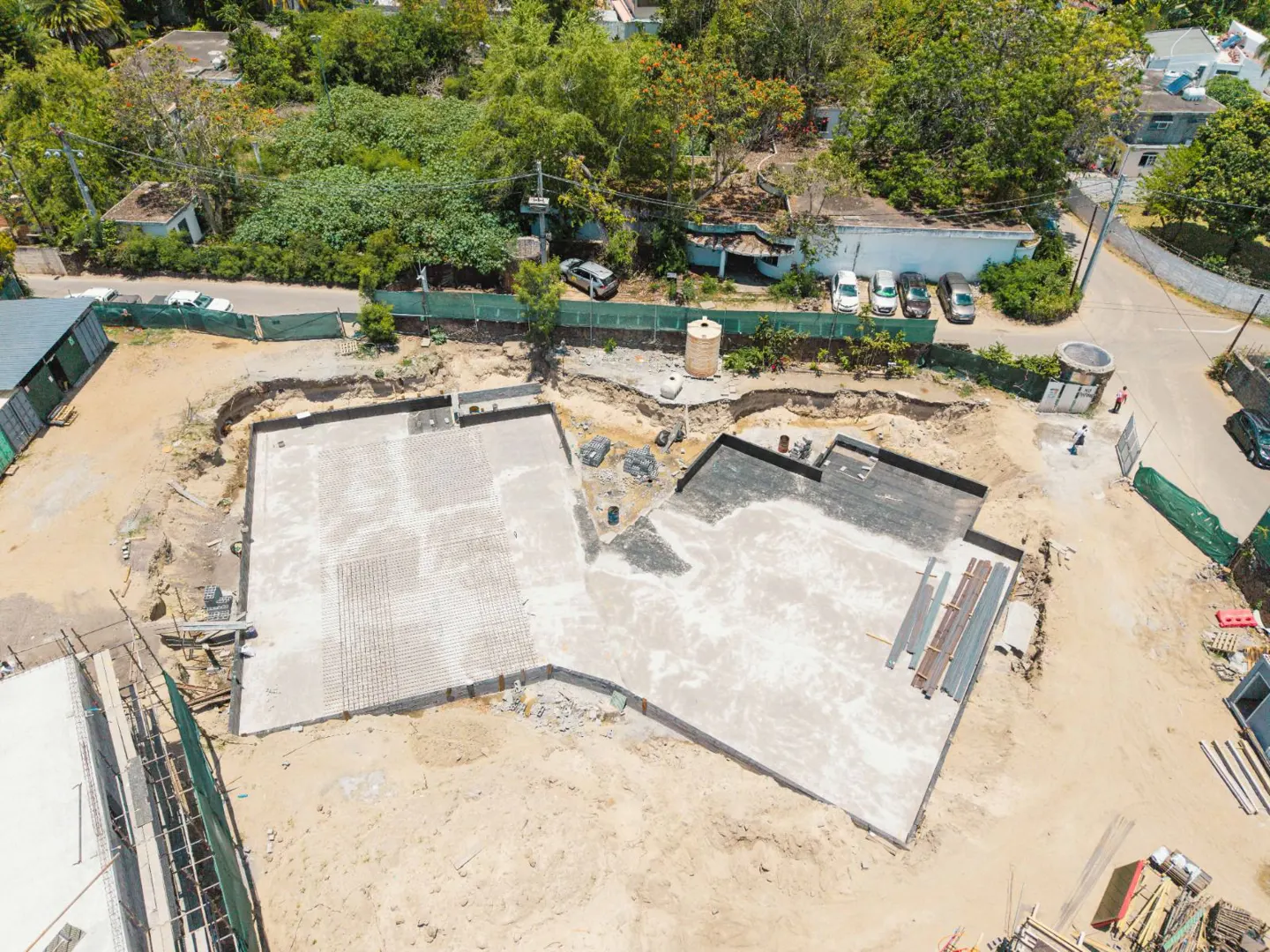 Aerial view of a construction site with a concrete foundation, surrounded by sand, green trees, and buildings in the background.