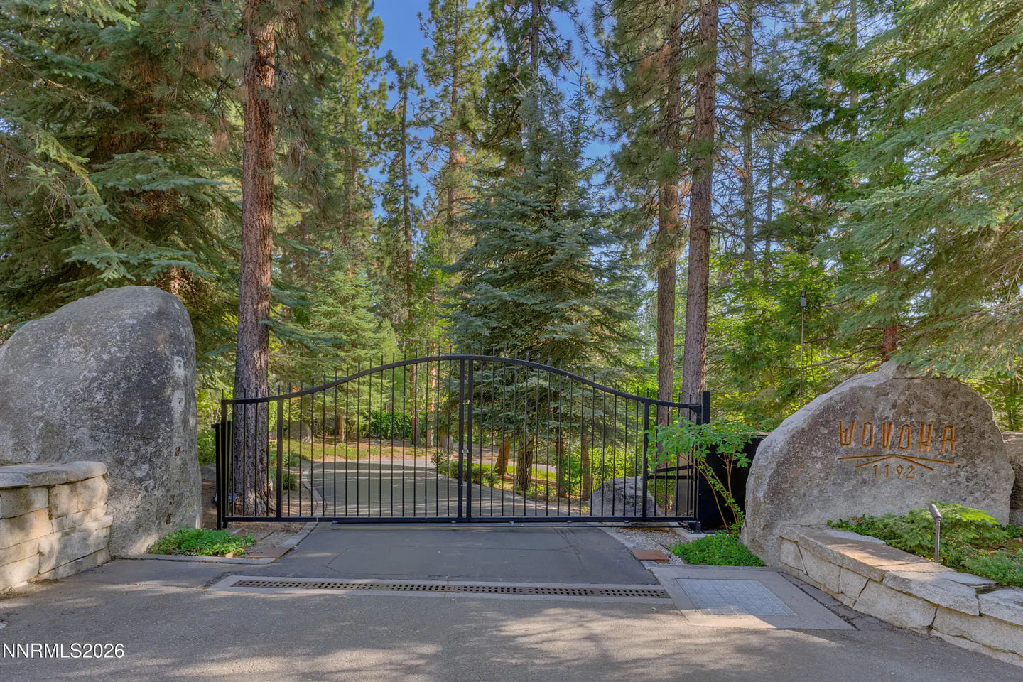 Gated entrance to a property. Black metal gate between two large boulders, surrounded by tall green trees. "WONOHA 1192" is carved into the right boulder.