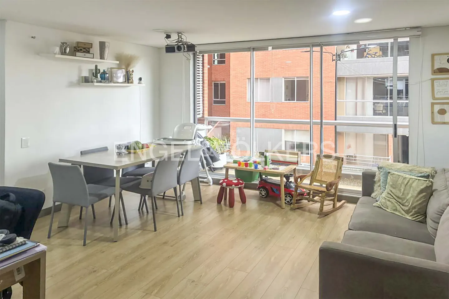 Bright living room with wood floors, gray sofa and chairs, dining table, and children's play area by a large window.