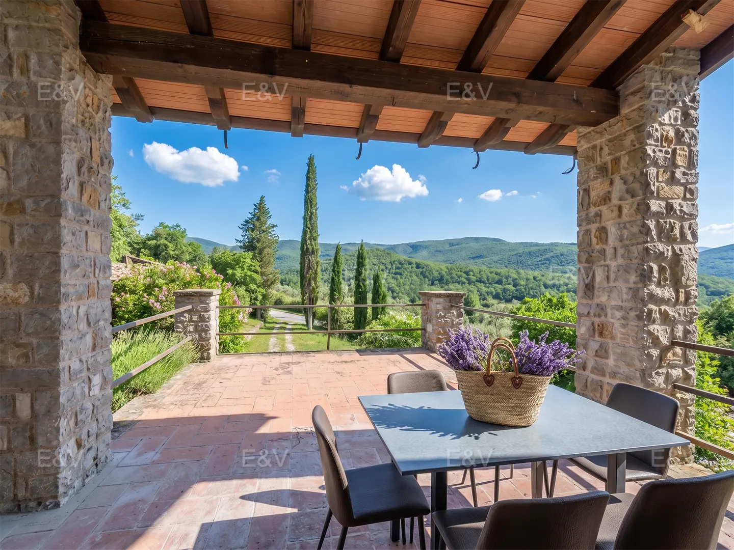 Covered patio with stone pillars, a table with chairs, and a basket of lavender overlooking a green valley on a sunny day.