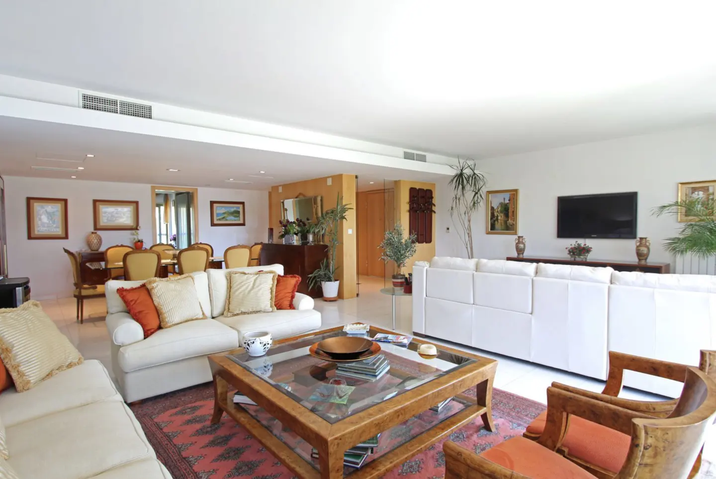 Bright living room with white sofas, wood chairs, and a glass-topped coffee table on a red patterned rug. Dining area visible in the background.