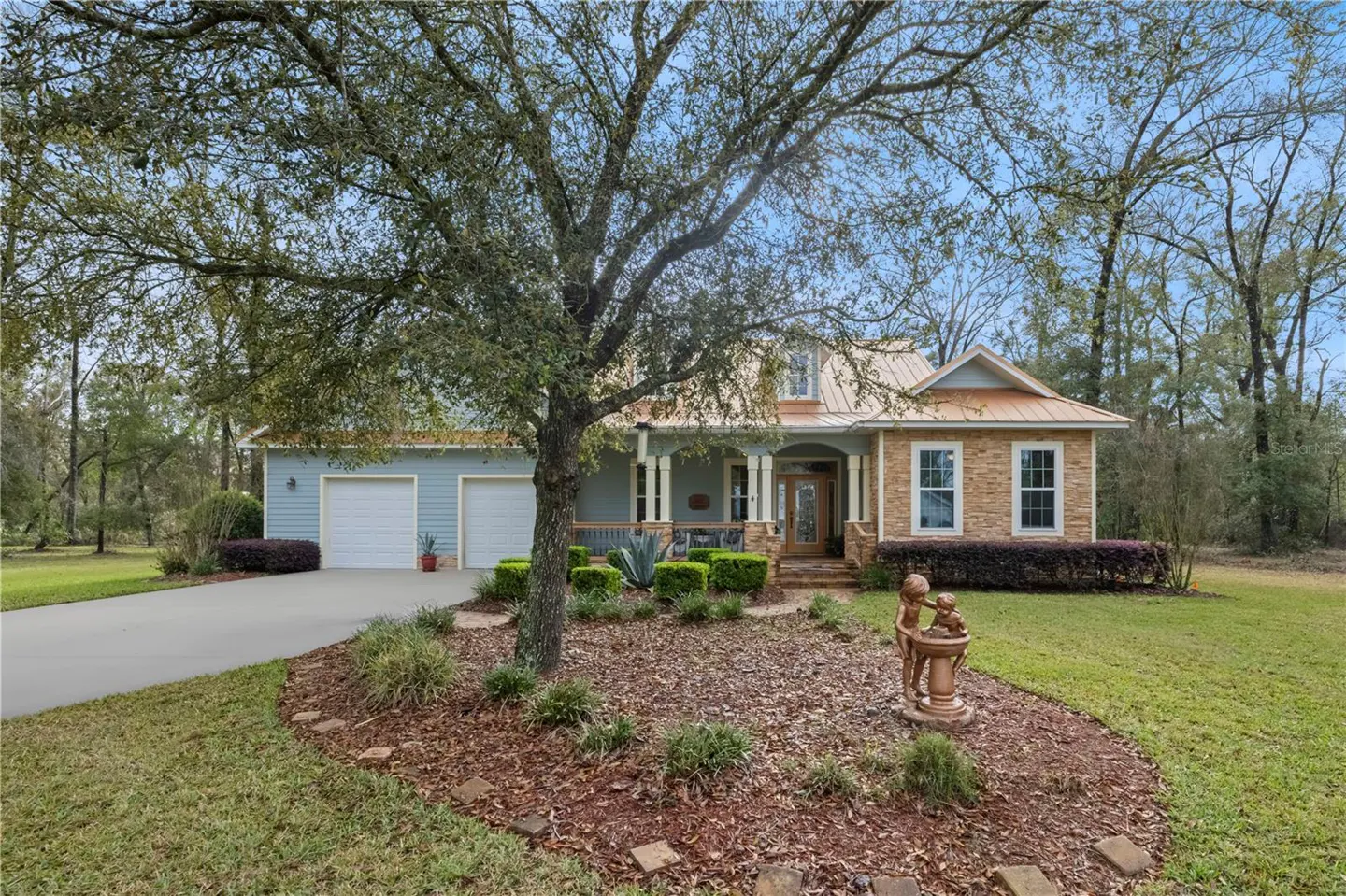 Two-story house with a light blue exterior, tan brick accents, and a metal roof. A large tree is in the front yard.