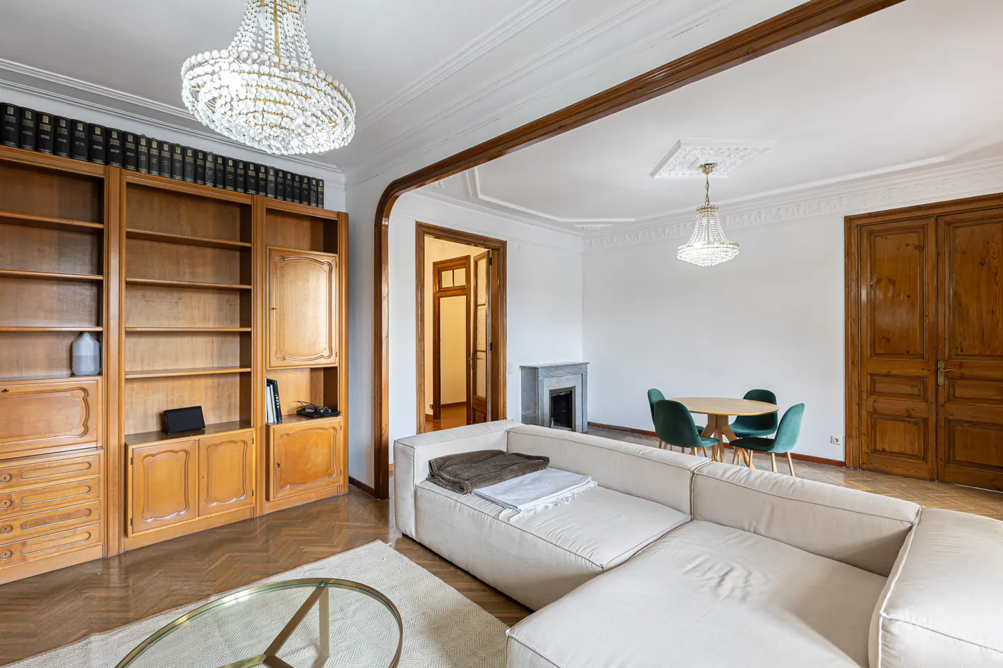 Bright living room with hardwood floors, white walls, and a large white sectional sofa. A wooden bookcase, dining table with green chairs, and crystal chandeliers are also visible.